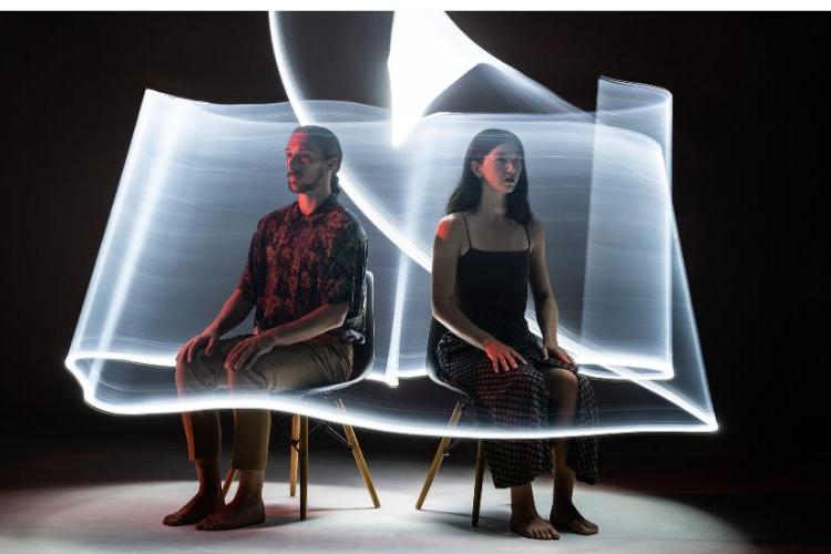 2 dancers seated in chairs are wrapped by a white ribbon of light.